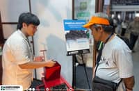 A DOST-FPRDI representative demonstrates a red charcoal briquetting machine to a visitor during the 2026 Regional Science, Technology, and Innovation Week at the Quezon Convention Center in Lucena City, Quezon. A display featuring charcoal briquettes is visible in the background.