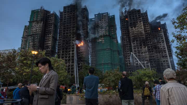 (FILES) People watch as smoke rises from apartments after a major fire swept through several blocks at the Wang Fuk Court residential estate in Hong Kong's Tai Po district on November 27, 2025. Hong Kong leader John Lee on October 29, 2026 apologised over the deadly fire that killed 168 people last year as the city's housing minister resigned after a report found the government "partially responsible" for the disaster.